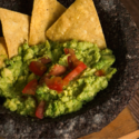 A close-up image of guacamole with tomato chunks served in a stone molcajete with tortilla chips arranged around the edges.