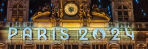 Illuminated signage "PARIS 2024" on a historically styled building facade with a clock, sculptures, and hanging flags at night.
