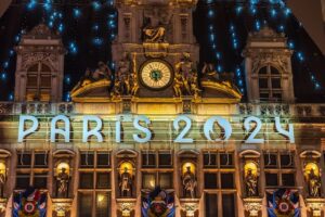 Illuminated signage "PARIS 2024" on a historically styled building facade with a clock, sculptures, and hanging flags at night.